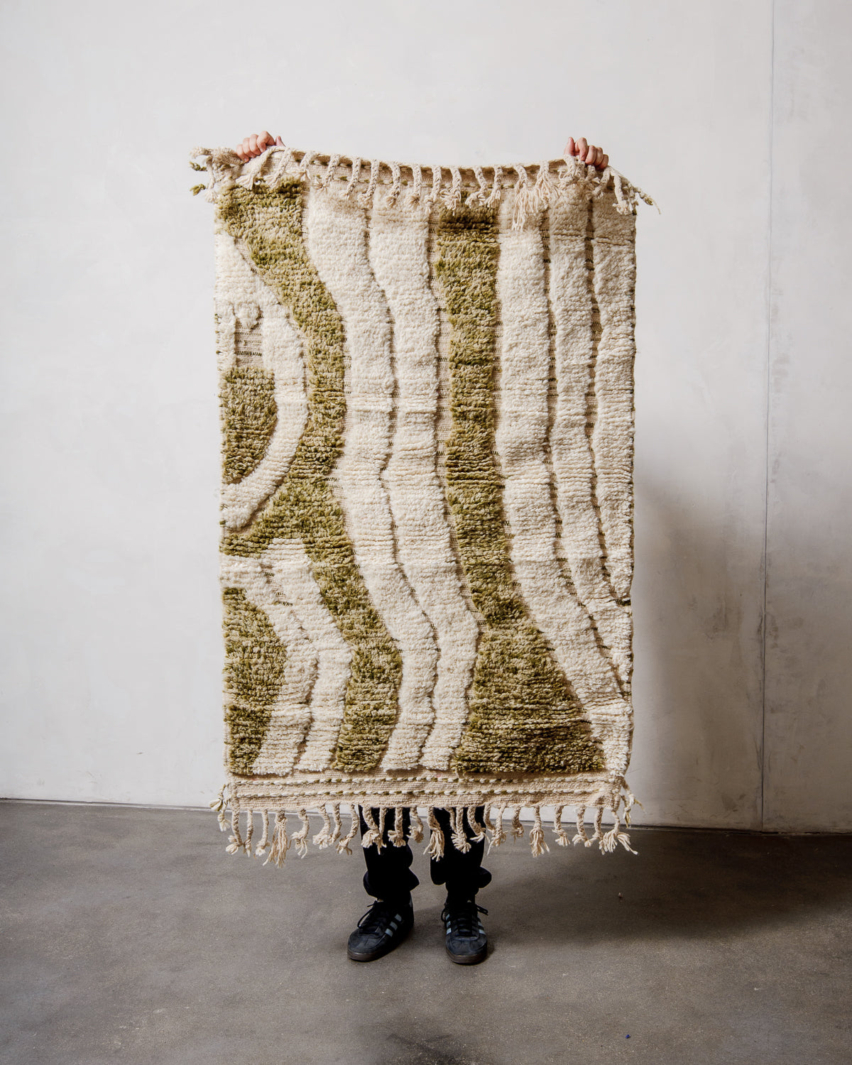 Small wool Berberlin Beniourain rug in cream featuring speckled khaki green, being held up in front of a white wall.